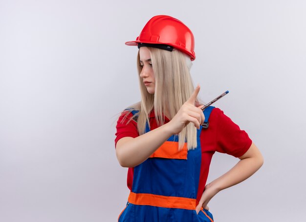 Giovane ragazza bionda del costruttore dell'ingegnere in uniforme che tiene la penna e che indica al lato destro con un'altra mano sulla vita guardando il lato sinistro sul muro bianco isolato con lo spazio della copia