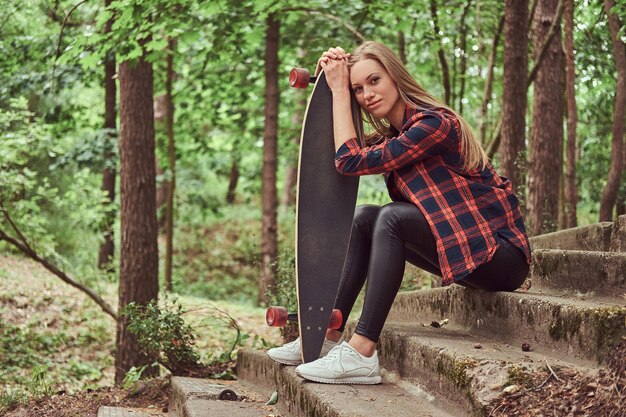 Giovane ragazza bionda hipster che riposa in un parco, dopo aver cavalcato su uno skateboard.