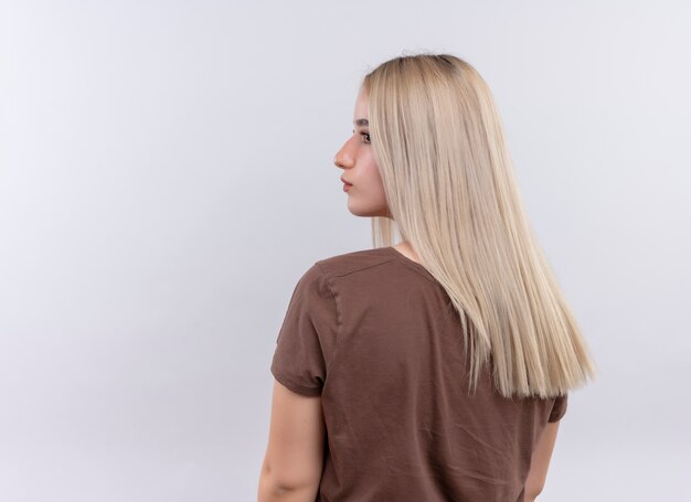 Giovane ragazza bionda in piedi dietro la vista guardando il lato sinistro sul muro bianco isolato con lo spazio della copia