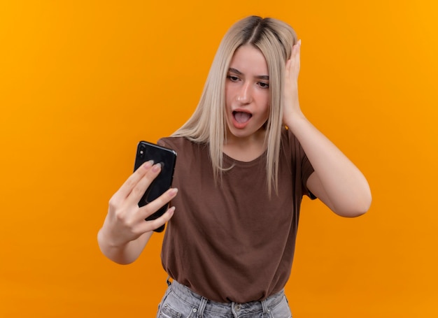 Giovane ragazza bionda sorpresa che tiene il telefono cellulare guardandolo con la mano sulla testa sulla parete arancione isolata con lo spazio della copia