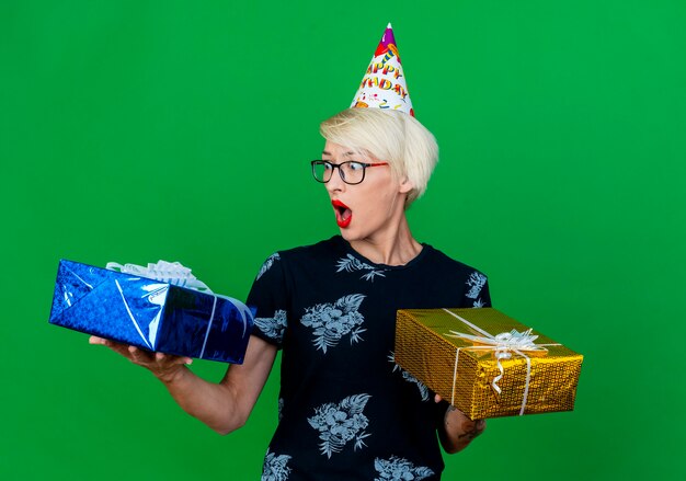 Giovane ragazza bionda sorpresa del partito con gli occhiali e cappello di compleanno che tengono i contenitori di regalo che esaminano uno di loro isolato su priorità bassa verde