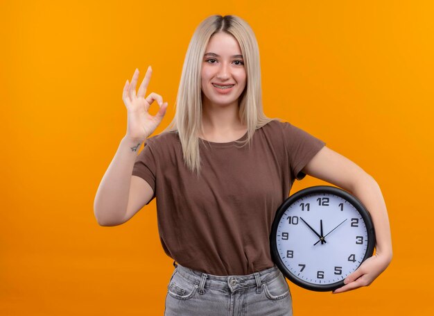 Giovane ragazza bionda sorridente in parentesi graffe dentali che tengono orologio che fa segno giusto sullo spazio arancione isolato con lo spazio della copia