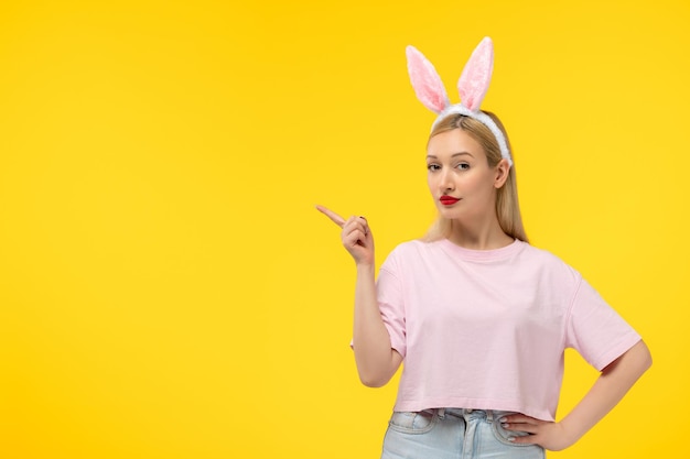 Giovane ragazza bionda sveglia di Pasqua che indossa le orecchie rosa del coniglietto che puntano con il dito a sinistra