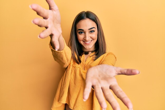 Giovane ragazza bruna che indossa abiti casual guardando la telecamera sorridente con le braccia aperte per l'abbraccio. espressione allegra che abbraccia la felicità.