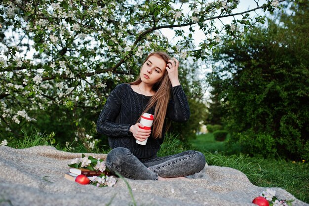 Giovane ragazza bruna in jeans seduta sul plaid contro l'albero in fiore di primavera e tenendo il thermos del tè a portata di mano