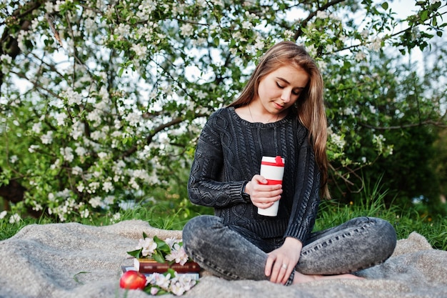 Giovane ragazza bruna in jeans seduta sul plaid contro l'albero in fiore di primavera e tenendo il thermos del tè a portata di mano