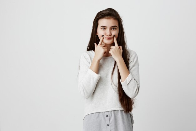 giovane ragazza bruna sconvolta adolescente facendo un sorriso falso con le dita che allungano gli angoli della bocca. Ritratto di donna che cerca di rimanere positivo