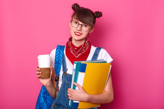 giovane ragazza bruna snella in maglietta bianca casual, tuta e bandana sul collo, con in mano una tazza di caffè e una cartella con documenti, in posa isolato su roseo. Concetto di giovane.