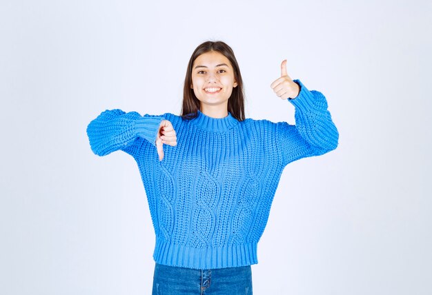 Giovane ragazza castana in maglione blu che dà i pollici giù e su su bianco.