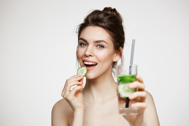 Giovane ragazza castana positiva che sorride esaminando macchina fotografica che mangia la fetta del cetriolo che tiene bicchiere d'acqua sopra fondo bianco. Bellezza e salute.