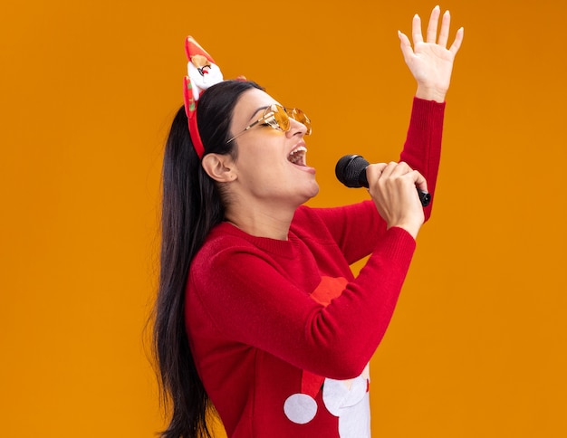 giovane ragazza caucasica che indossa la fascia e il maglione di babbo natale con gli occhiali in piedi nella vista di profilo tenendo il microfono vicino alla bocca cantando con gli occhi chiusi alzando la mano isolata sul muro arancione con spazio di copia