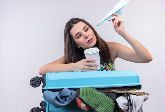 Giovane ragazza caucasica del viaggiatore che tiene la tazza di caffè di plastica di viaggio e l'aereo di carta su fondo bianco isolato