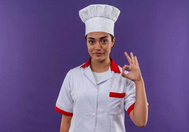 Giovane ragazza caucasica felice del cuoco in segno giusto della mano di gesti uniformi del cuoco unico isolato sulla parete viola con lo spazio della copia
