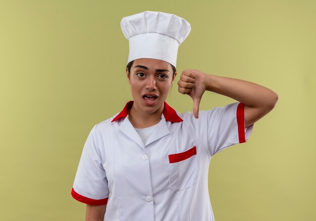 Giovane ragazza caucasica infastidita del cuoco in uniforme del cuoco unico i pollici giù isolati su fondo verde con lo spazio della copia