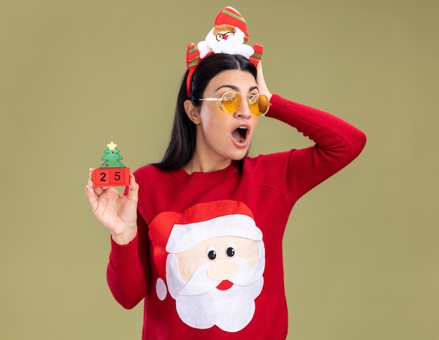 Giovane ragazza caucasica interessata che indossa la fascia e il maglione di Babbo Natale con gli occhiali che tengono il giocattolo dell'albero di Natale con la data che tiene la mano sulla testa guardando il lato isolato sulla parete verde oliva