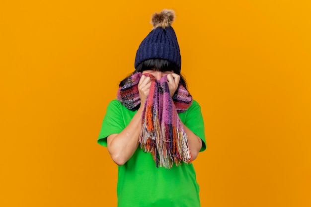 Giovane ragazza caucasica malata che indossa il cappello e la sciarpa di inverno che esaminano la sciarpa della tenuta della macchina fotografica sulla faccia isolata su fondo arancio con lo spazio della copia