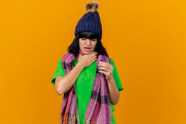 Giovane ragazza caucasica malata dolorante che indossa cappello invernale e sciarpa tenendo le capsule tenendo la mano sulla gola con gli occhi chiusi isolati su sfondo arancione con spazio di copia