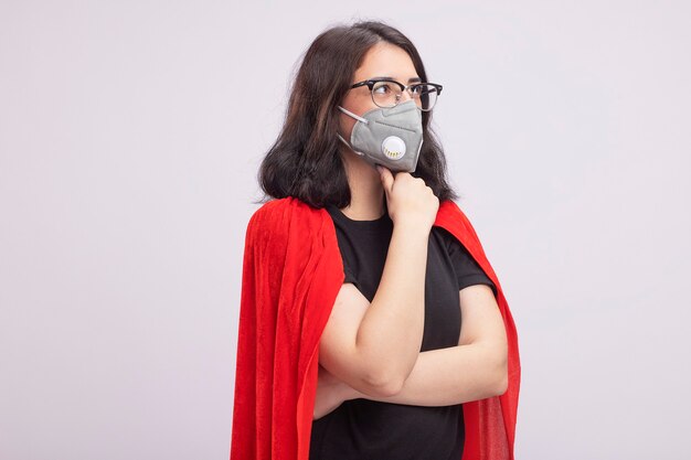 Giovane ragazza caucasica premurosa del supereroe in mantello rosso con gli occhiali e maschera protettiva che tocca il mento guardando il lato isolato sul muro bianco con spazio di copia