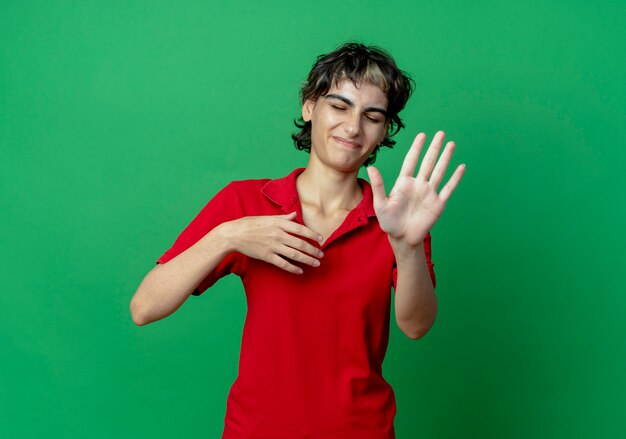 Giovane ragazza caucasica scontenta con un taglio di capelli da folletto che tiene la mano in aria e fa un gesto di no con gli occhi chiusi closed