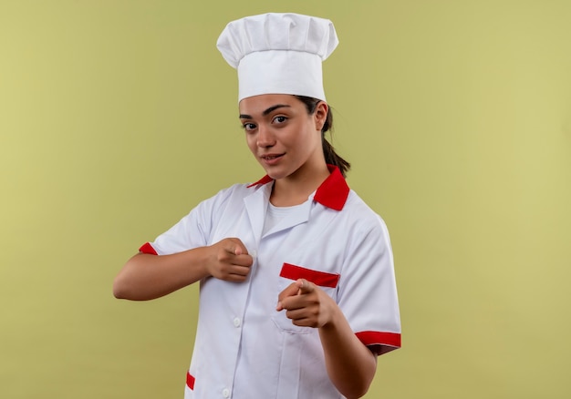 Giovane ragazza caucasica sicura del cuoco nei punti dell'uniforme del cuoco unico con entrambe le dita isolate sulla parete verde con lo spazio della copia