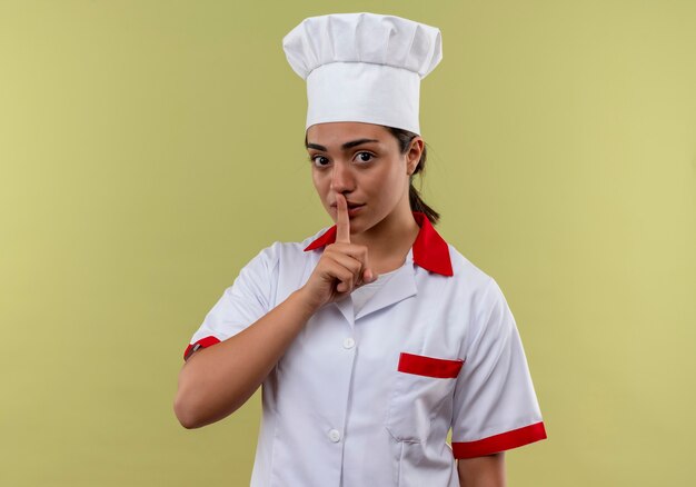 Giovane ragazza caucasica sicura del cuoco nel segno della mano di silenzio di gesti uniformi dello chef isolato sulla parete verde con lo spazio della copia