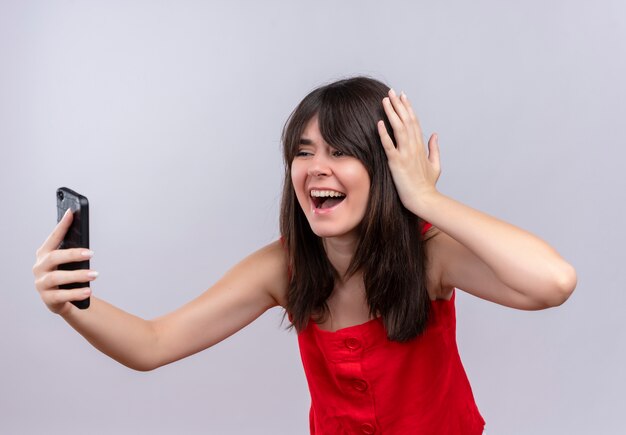 Giovane ragazza caucasica sorpresa che tiene telefono e che tiene testa che esamina telefono su fondo bianco isolato