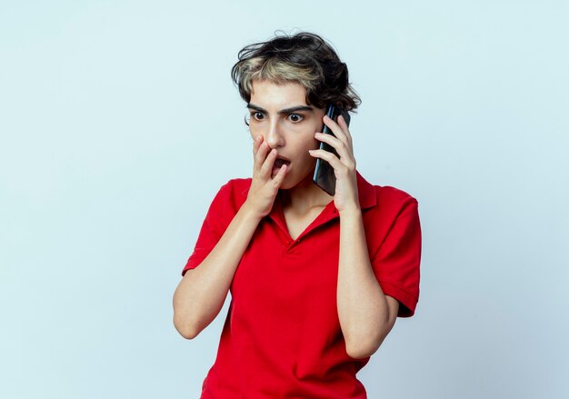 Giovane ragazza caucasica sorpresa con taglio di capelli del folletto che parla sul telefono che mette la mano sulla bocca che osserva giù isolata su fondo bianco con lo spazio della copia