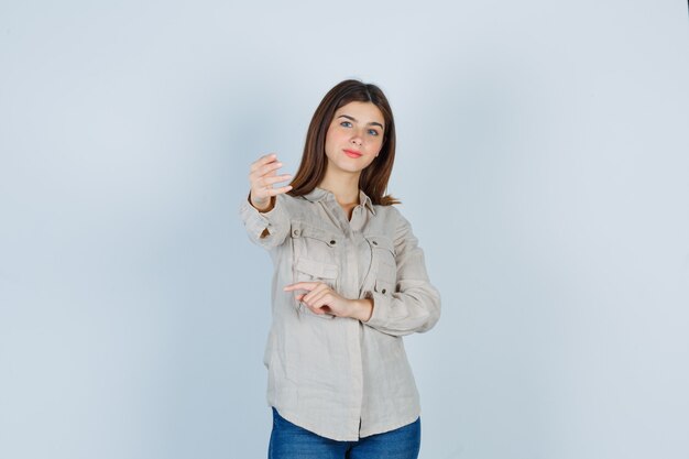 Giovane ragazza che allunga la mano verso, puntando a sinistra con il dito indice in camicia beige, jeans e sembra felice. vista frontale.