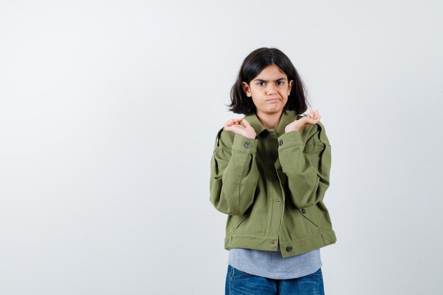 Giovane ragazza che allunga le mani in modo interrogativo in maglione grigio, giacca color kaki, pantaloni di jeans e sembra perplessa, vista frontale.