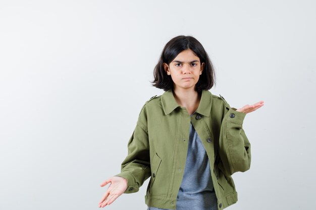 Giovane ragazza che allunga le mani in modo interrogativo in maglione grigio, giacca kaki, pantaloni jeans e sembra sconcertata, vista frontale.