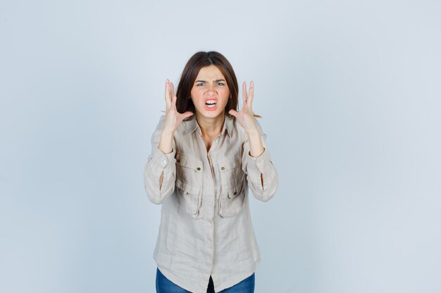 Giovane ragazza che alza le mani vicino alla testa in modo arrabbiato in camicia beige, jeans e sembra agitata. vista frontale.