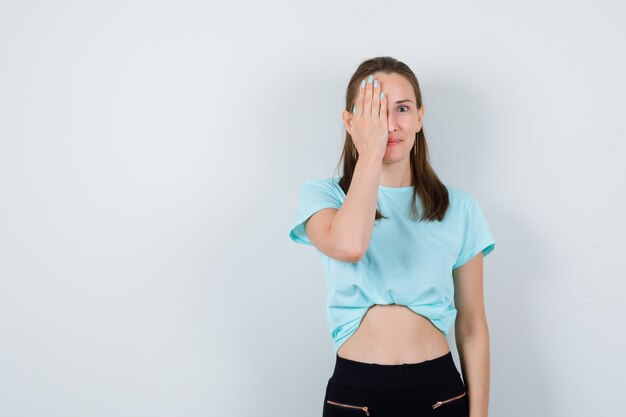 Giovane ragazza che copre il viso con il palmo in maglietta turchese, pantaloni e sguardo curioso, vista frontale.