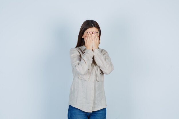 Giovane ragazza che copre il viso con le mani in camicia beige, jeans e si vergogna. vista frontale.