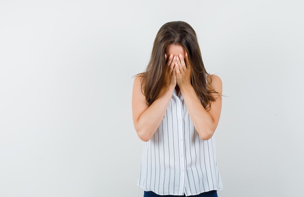 Giovane ragazza che copre il viso con le mani in t-shirt, jeans e sembra stanco, vista frontale.