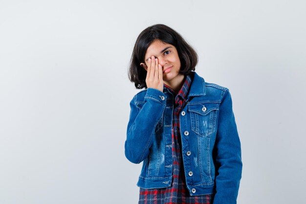 Giovane ragazza che copre l'occhio con la mano in camicia a quadri e giacca di jeans e sembra seria.