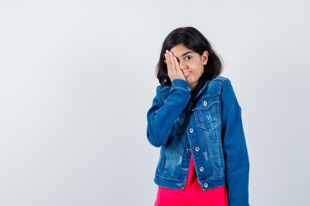 Giovane ragazza che copre l'occhio con la mano in maglietta rossa e giacca di jeans e sembra felice, vista frontale.