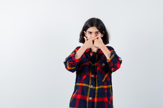 Giovane ragazza che copre la bocca con le mani, indicando direzioni diverse in camicia a quadri e guardando seria, vista frontale.