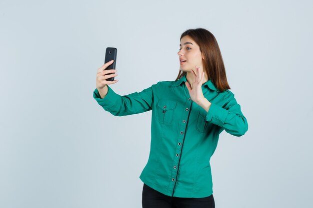 Giovane ragazza che fa videochiamata, agitando la mano per dire ciao in camicetta verde, pantaloni neri e guardando felice, vista frontale.