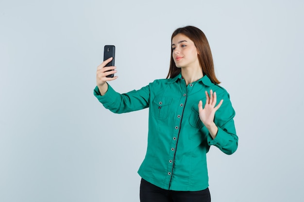 Giovane ragazza che fa videochiamata, agitando la mano per salutare in camicetta verde, pantaloni neri e guardando allegro, vista frontale.