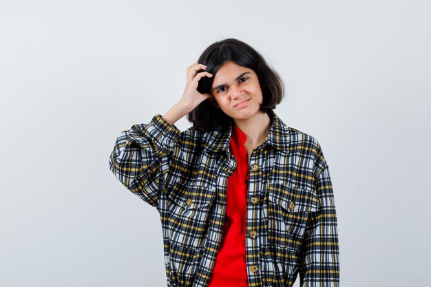 Giovane ragazza che graffia la testa in camicia a quadri e maglietta rossa e sembra seria. vista frontale.