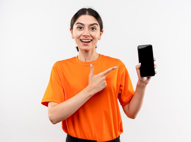 Giovane ragazza che indossa la maglietta arancione che mostra lo smartphone che punta con il dito indice a sorridere allegramente in piedi sopra il muro bianco