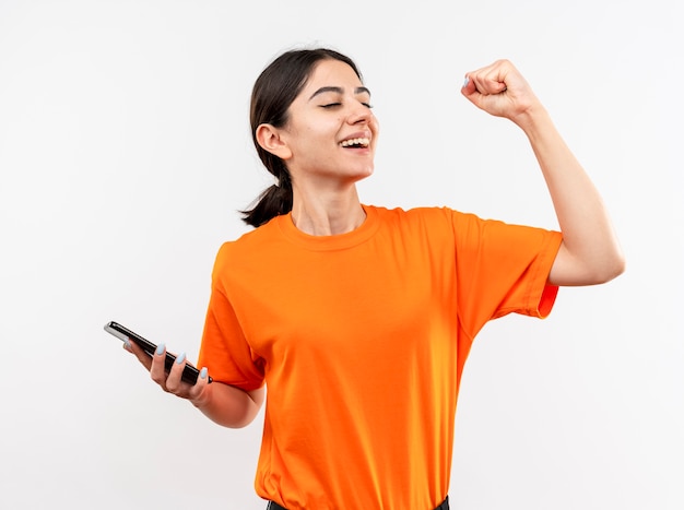 Giovane ragazza che indossa la maglietta arancione che tiene smartphone pugno di serraggio felice ed eccitato sorridendo allegramente rallegrandosi del suo successo in piedi sopra il muro bianco