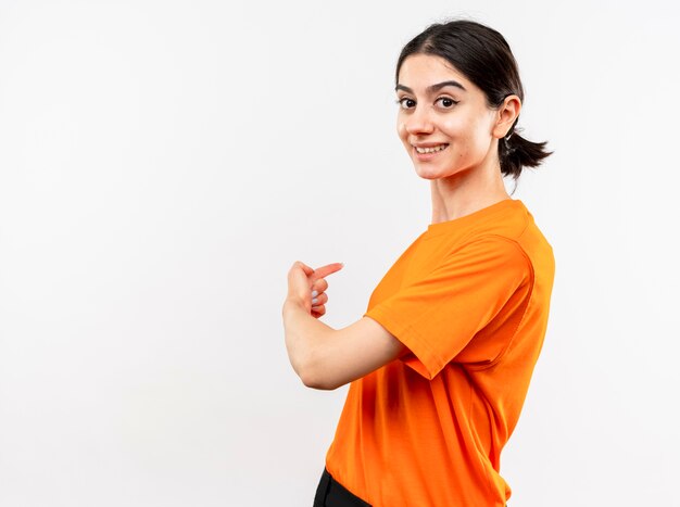 Giovane ragazza che indossa la maglietta arancione con il sorriso sul viso rivolto indietro con il dito in piedi sul muro bianco