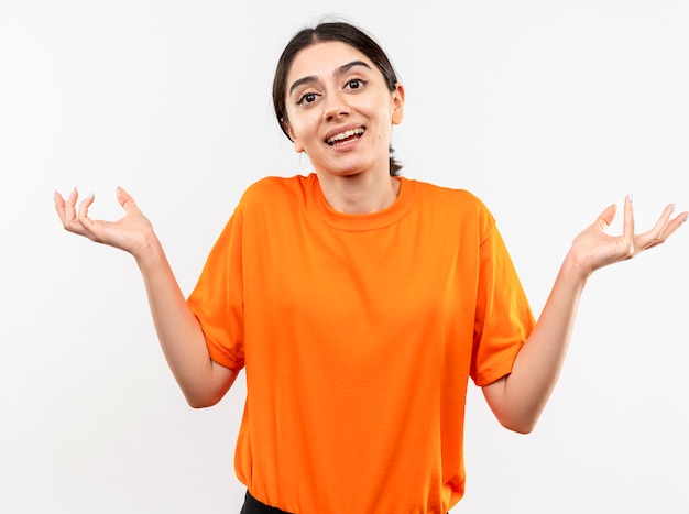 Giovane ragazza che indossa la maglietta arancione essendo confusa e incerta sorridente diffondendo le braccia ai lati avendo dubbi in piedi sul muro bianco