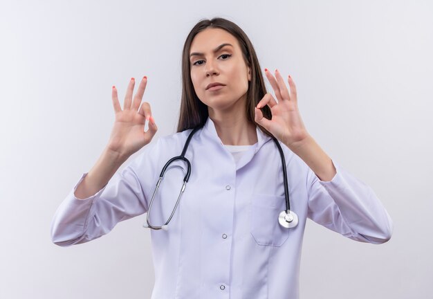 giovane ragazza che indossa stetoscopio abito medico che mostra okey gesto sul muro bianco isolato