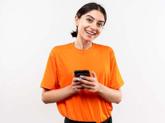 Giovane ragazza che indossa t-shirt arancione tenendo lo smartphone sorridente con la faccia felice in piedi sopra il muro bianco