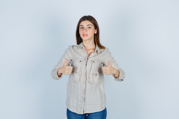 Giovane ragazza che mostra doppio pollice in alto in camicia beige, jeans e guardando seria, vista frontale.