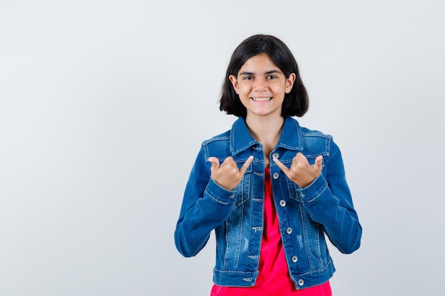 Giovane ragazza che mostra gesti di pistola, sorridente in maglietta rossa e giacca di jeans e sembra carina. vista frontale.