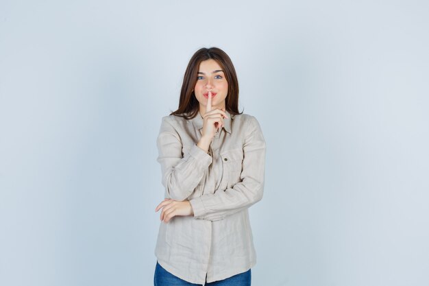 Giovane ragazza che mostra gesto di silenzio in camicia beige, jeans e sguardo serio. vista frontale.