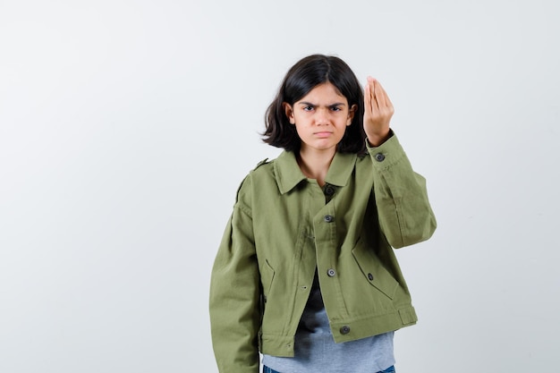 Giovane ragazza che mostra gesto italiano in maglione grigio, giacca kaki, pantaloni jeans e sembra arrabbiata. vista frontale.